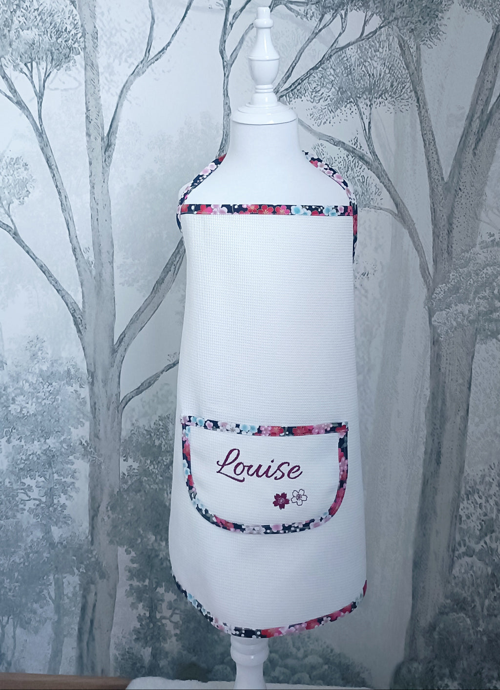 Tablier de cuisine enfant en tissu nid d'abeille blanc présenté sur mannequin. Poche avant brodée avec le prénom Louise et finitions biais fleuri.