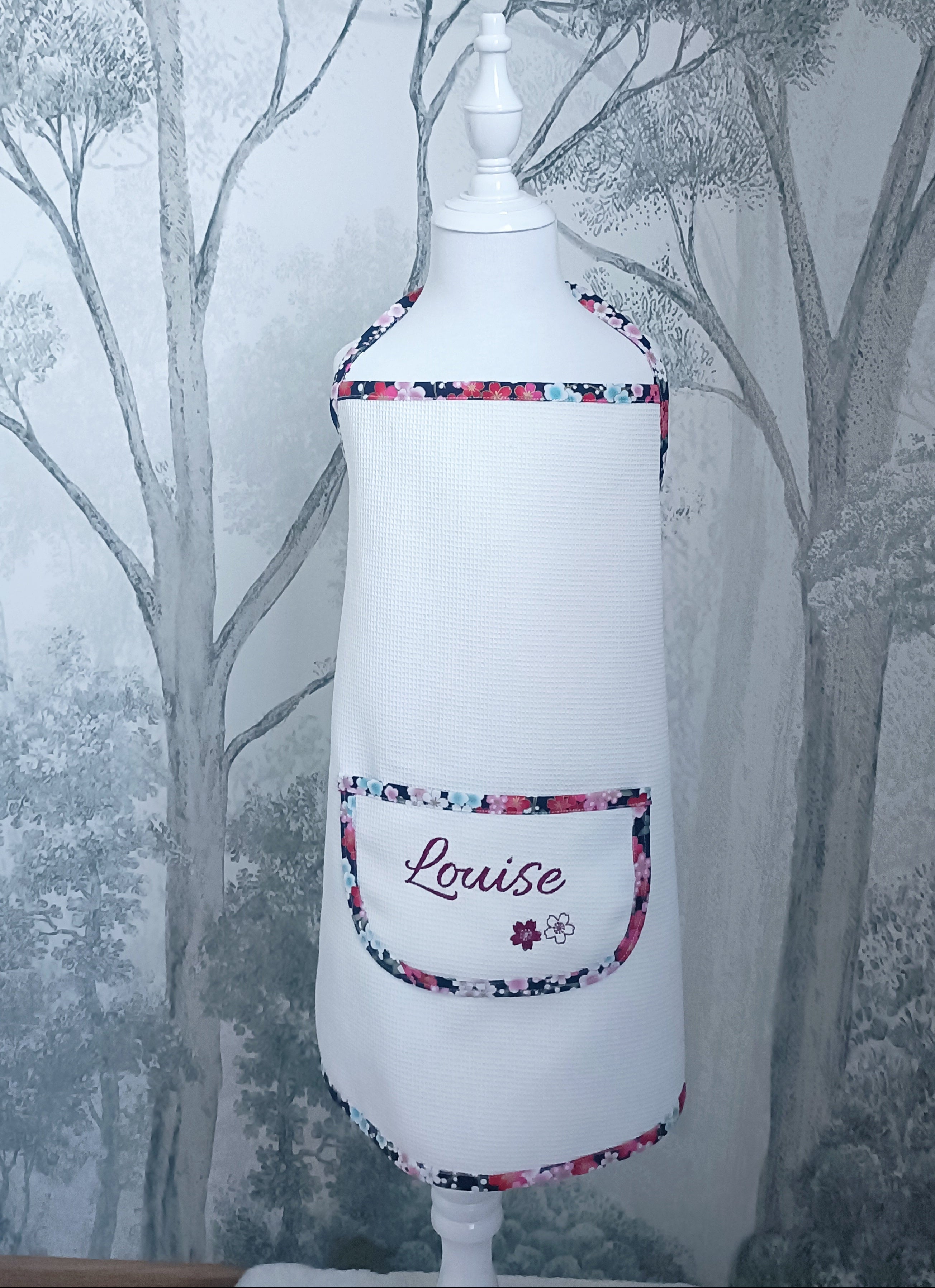 Tablier de cuisine enfant en tissu nid d'abeille blanc présenté sur mannequin. Poche avant brodée avec le prénom Louise et finitions biais fleuri.