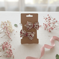 Duo de barrettes à nœud floral rouge et bordeaux présenté dans une ambiance douce avec ruban rose et fleurs séchées. Accessoire de coiffure élégant pour fille.