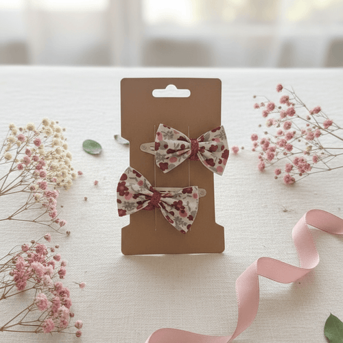 Duo de barrettes à nœud floral rouge et bordeaux présenté dans une ambiance douce avec ruban rose et fleurs séchées. Accessoire de coiffure élégant pour fille.