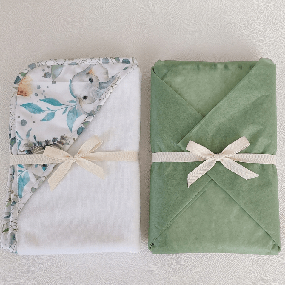 Deux capes de bain bébé faites main pliées et nouées d'un ruban ivoire — motif safari aquarelle éléphant sur fond blanc et uni vert sauge, L'Atelier de Megh.