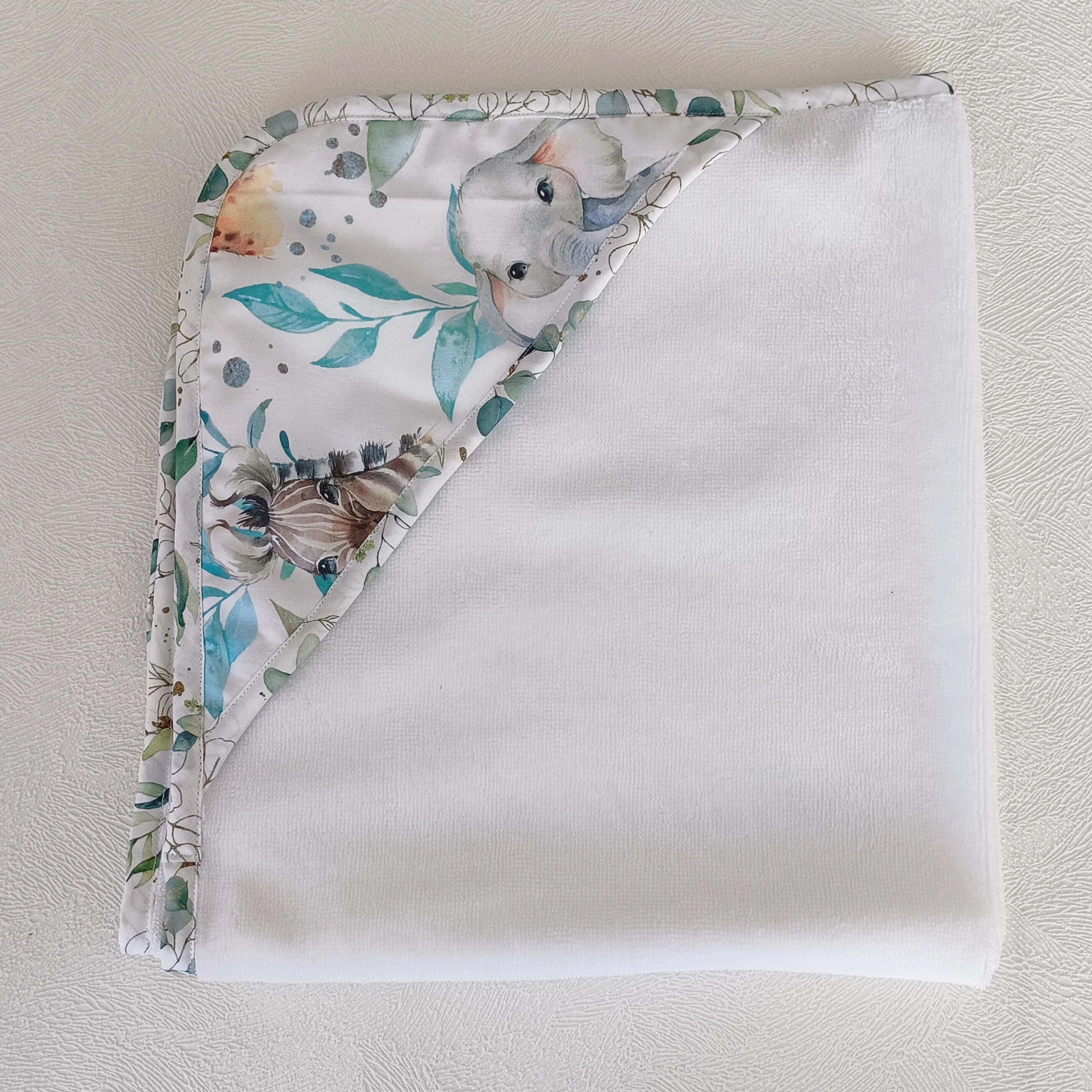 Cape de bain bébé personnalisable pliée vue de dessus, bordure motif éléphant et animaux safari aquarelle tons bleus et verts, éponge blanche, faite main L'Atelier de Megh