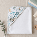Cape de bain bébé personnalisée avec broderie du prénom Timéo, motif éléphant et feuillage aquarelle, posée à plat sur lin naturel