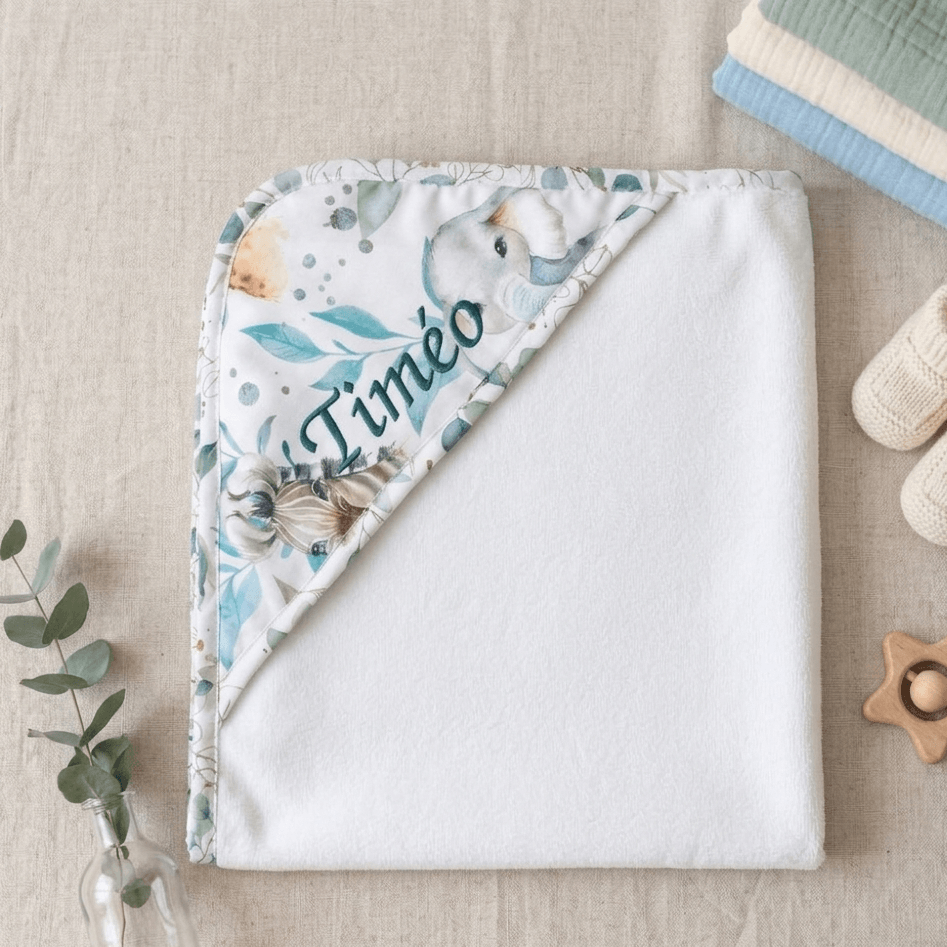 Cape de bain bébé personnalisée avec broderie du prénom Timéo, motif éléphant et feuillage aquarelle, posée à plat sur lin naturel