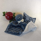 Doudou baleine bleu en tissu minky, avec pomme décorative et feuillage en arrière-plan.