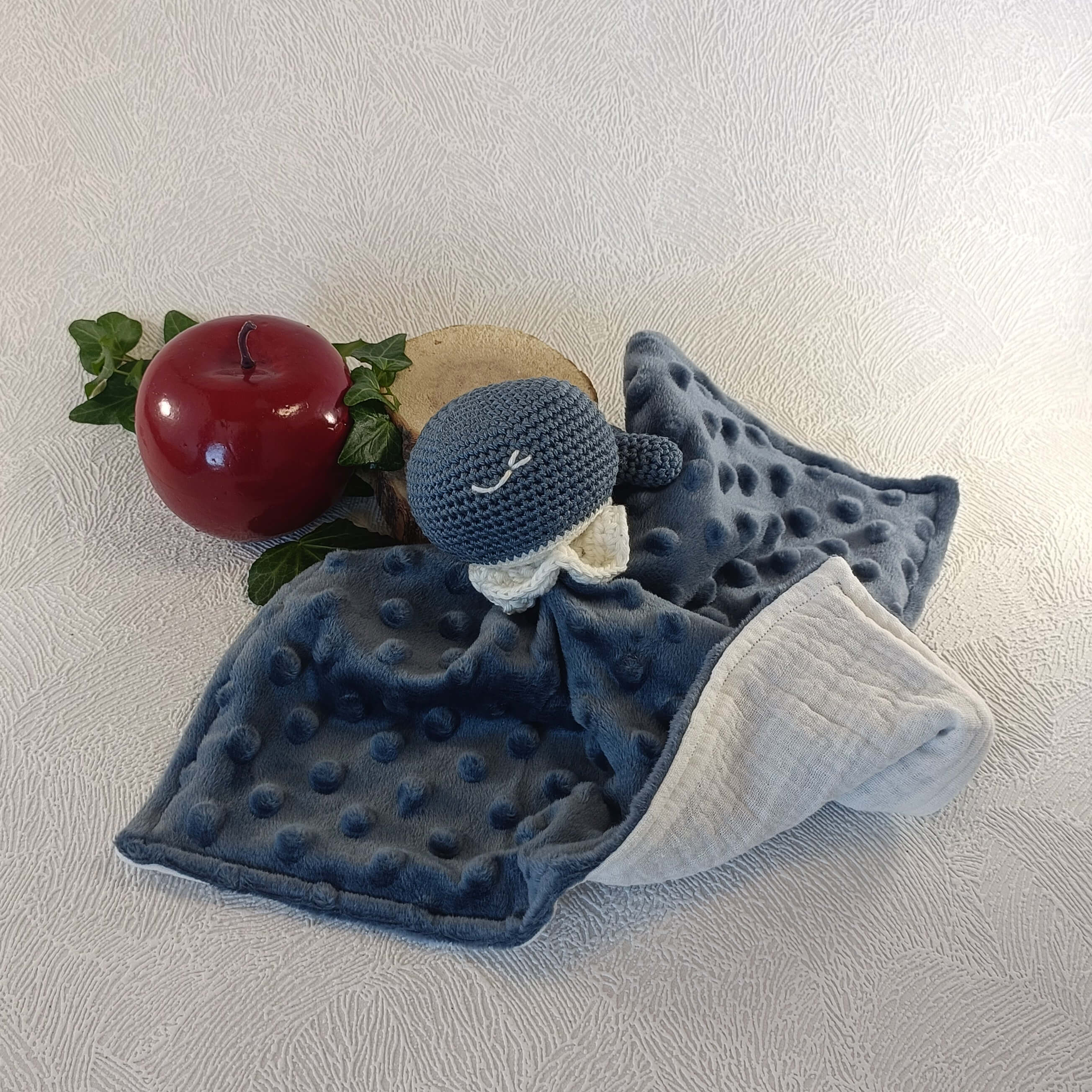 Doudou baleine bleu en tissu minky, avec pomme décorative et feuillage en arrière-plan.