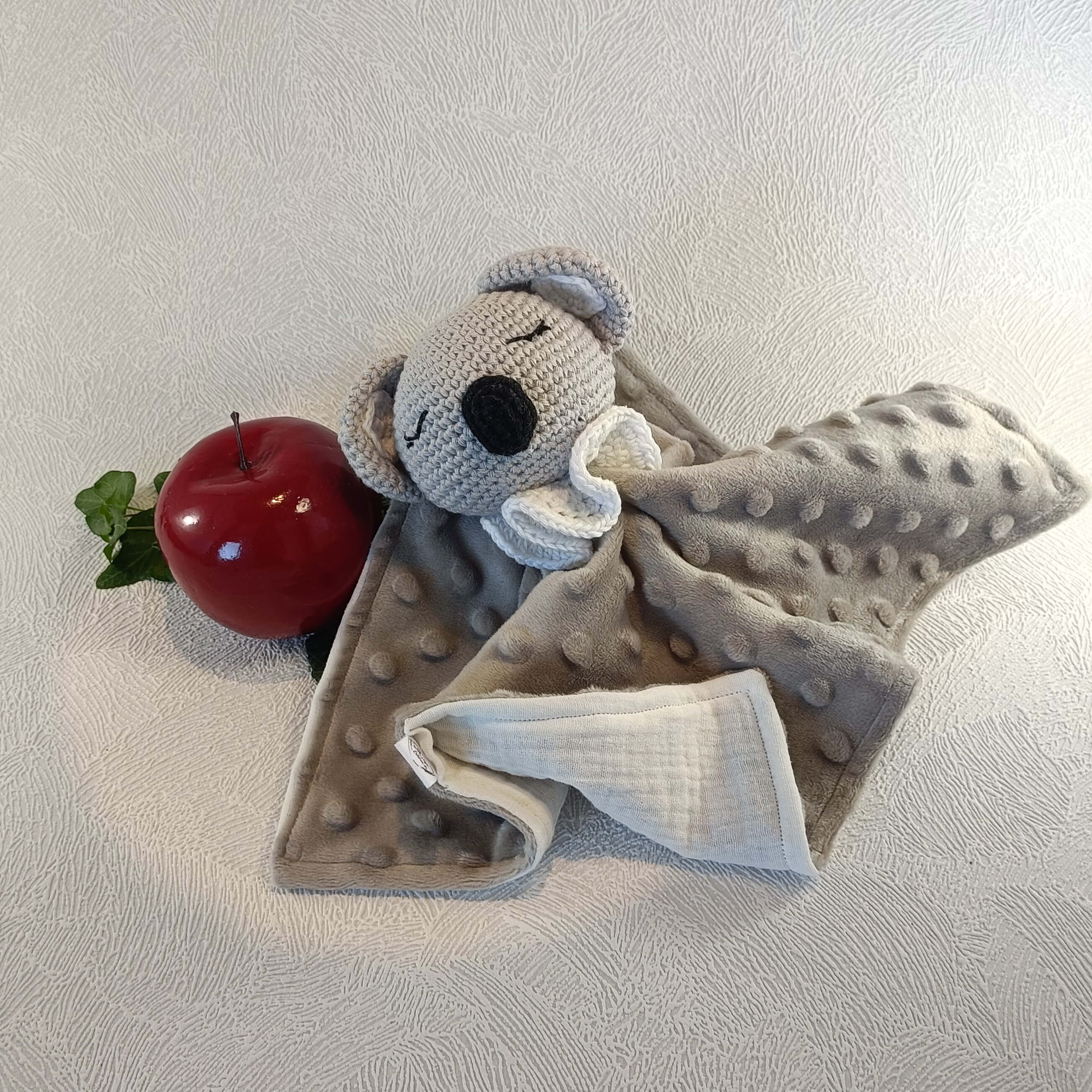 Doudou koala gris en tissu minky avec tête crochetée, à côté d'une pomme rouge