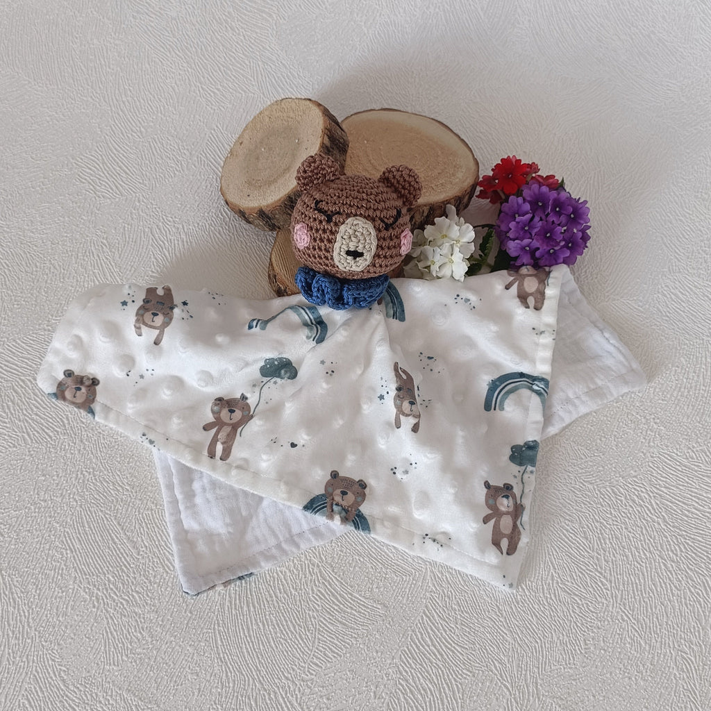 Doudou ours au crochet posé à plat sur lange double face — recto motif oursons avec arcs-en-ciel bleus, verso minky blanc à bulles