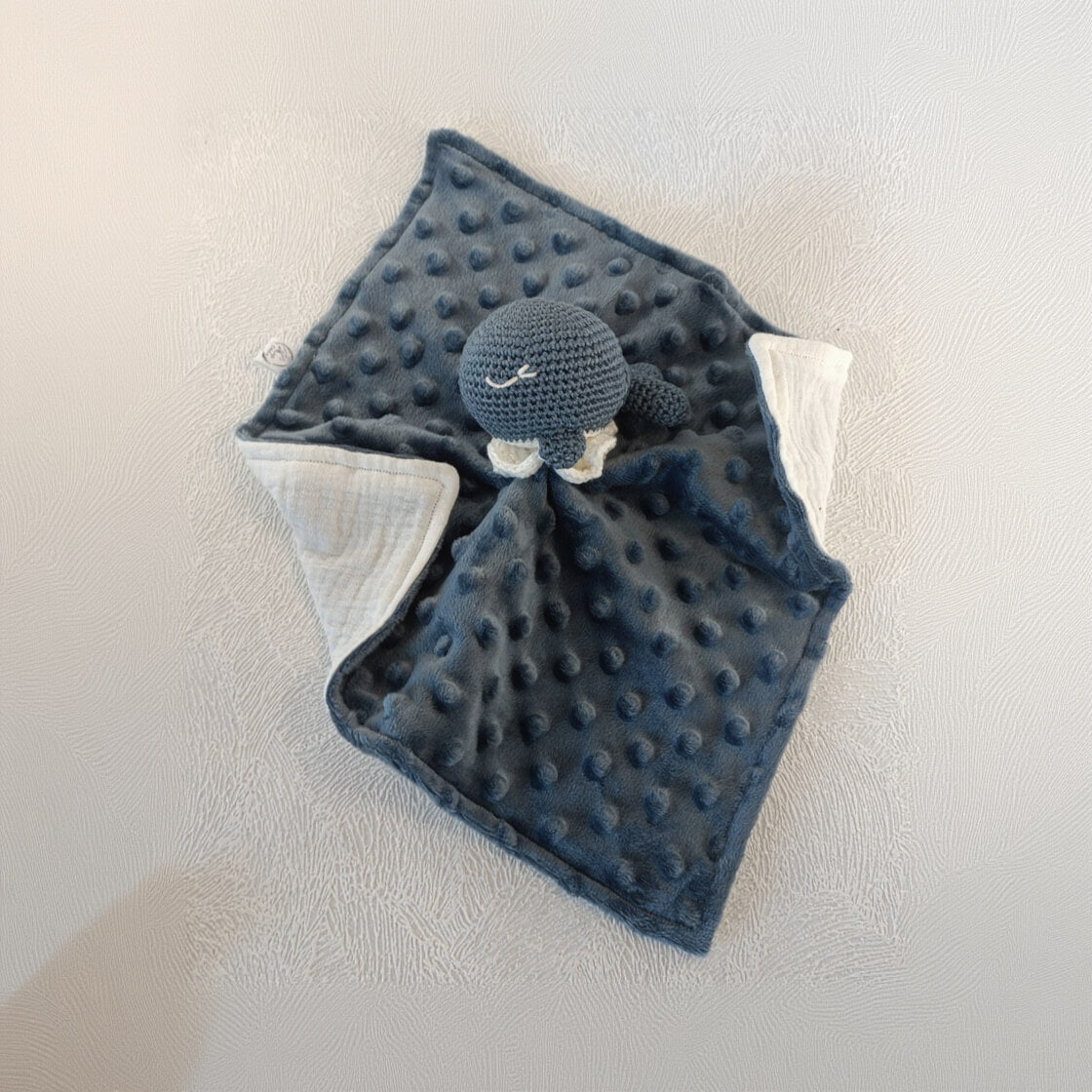 Doudou bébé bleu foncé à texture minky avec tête de baleine crochetée, posé sur table claire.