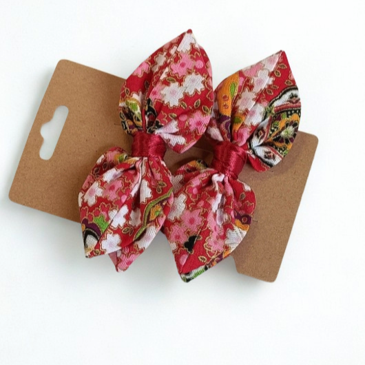 Duo de barrettes à cheveux forme nœud en tissu japonais rouge vif avec motif fleurs de cerisier (Sakura) roses et touches vertes. Centre du nœud en ruban rouge satiné.