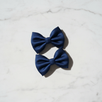 Vue de dessus de deux barrettes nœuds papillon bleu marine en tissu posées sur une surface en marbre blanc.