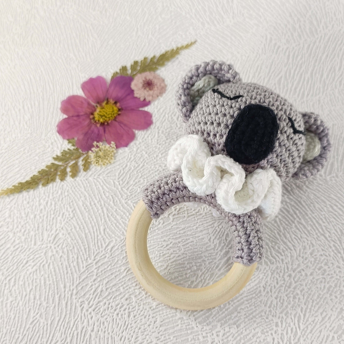 Hochet bébé koala au crochet avec anneau en bois, posé sur fond fleuri artisanal.