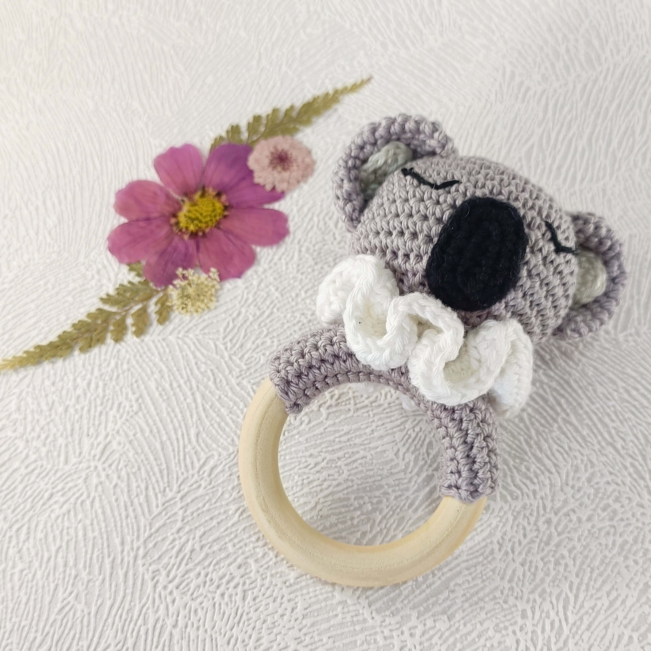 Hochet bébé koala au crochet avec anneau en bois, posé sur fond fleuri artisanal.