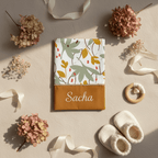 Mise en scène poétique d'un protège carnet de santé personnalisé Sacha au motif végétal automnal, entouré de chaussons de naissance en tricot et d'hortensias séchés.