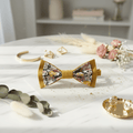Composition d'accessoires de mariage bohème : nœud papillon moutarde, bijoux dorés et fleurs séchées sur une table en marbre.