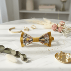 Composition d'accessoires de mariage bohème : nœud papillon moutarde, bijoux dorés et fleurs séchées sur une table en marbre.