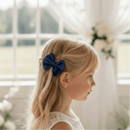 Portrait de profil d'une petite fille blonde portant un nœud papillon bleu marine dans les cheveux, vêtue d'une robe en dentelle blanche dans un décor de mariage lumineux.