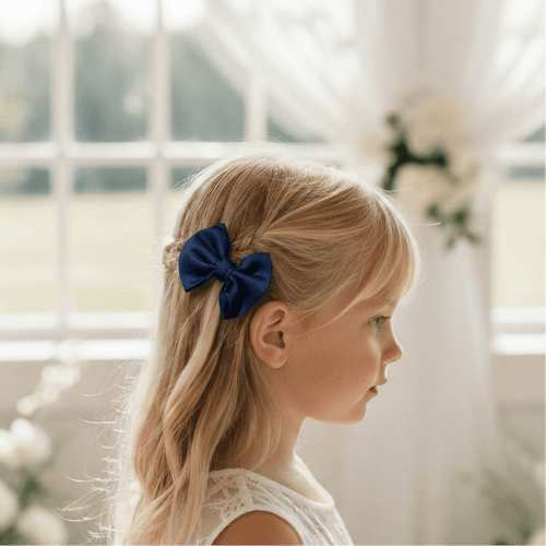 Portrait de profil d'une petite fille blonde portant un nœud papillon bleu marine dans les cheveux, vêtue d'une robe en dentelle blanche dans un décor de mariage lumineux.