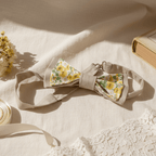 Nœud papillon fleuri posé sur un drapé en lin avec un livre ancien, de la dentelle et des fleurs séchées dans une ambiance lumineuse.