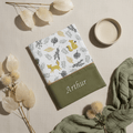 Mise en scène douce et naturelle présentant un protège carnet de santé brodé Arthur avec motif renards, entouré de fleurs séchées et d'un lange en gaze de coton vert kaki.