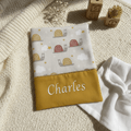 Protège carnet de santé personnalisé Charles brodé sur tissu jaune moutarde, avec motif escargots et étoiles, posé sur une couverture en tricot.