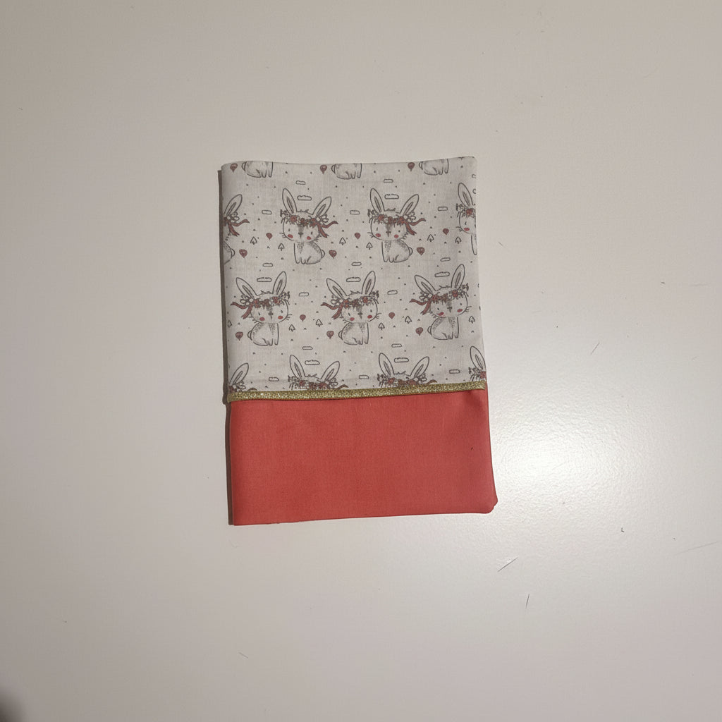 Vue de face d'un protège carnet de santé pour fille : tissu imprimé lapins mignons et partie inférieure en coton uni rose corail soulignée d'un liseré doré.