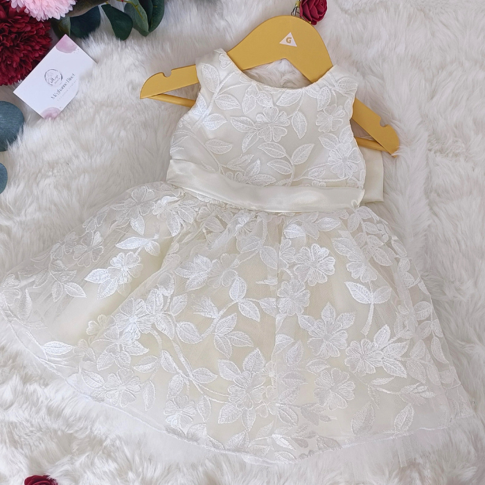 Robe de cérémonie blanche pour bébé fille présentée sur un cintre. Vue de face montrant les détails de la broderie florale et la ceinture satinée. Création artisanale.