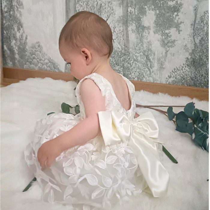 Bébé fille assise de dos portant la robe de cérémonie blanche. Gros plan sur le magnifique nœud en satin au dos et les détails de broderie florale sur le tulle.