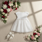 Robe de baptême fille blanche à haut crocheté et jupe en organza, présentée à plat avec chaussures et fleurs.