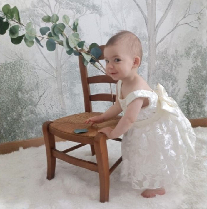 Bébé fille debout de profil portant la robe de cérémonie blanche. Mise en valeur du grand nœud en satin au dos et du volume de la jupe brodée.