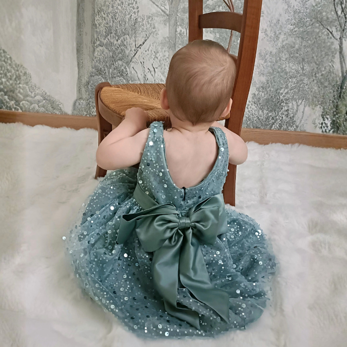 Bébé portant la robe de cérémonie vert sauge, vu de dos. Gros plan sur le grand nœud en satin et le tissu à sequins scintillants.