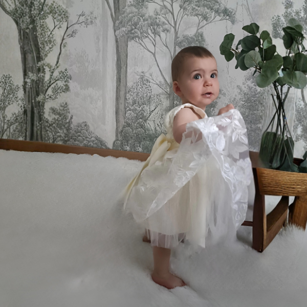 Bébé fille debout jouant avec sa robe de cérémonie blanche. Vue montrant la légèreté et la souplesse des jupons en tulle brodé.