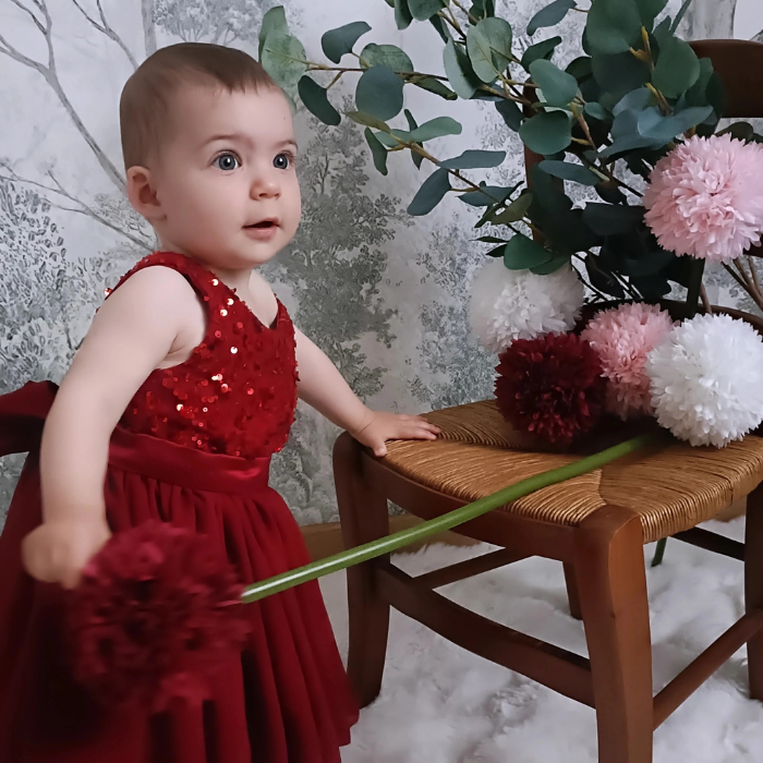 Bébé fille debout portant la robe de cérémonie rouge à paillettes. Vue en pied montrant le tombé fluide de la jupe en tulle. Tenue de fête élégante.