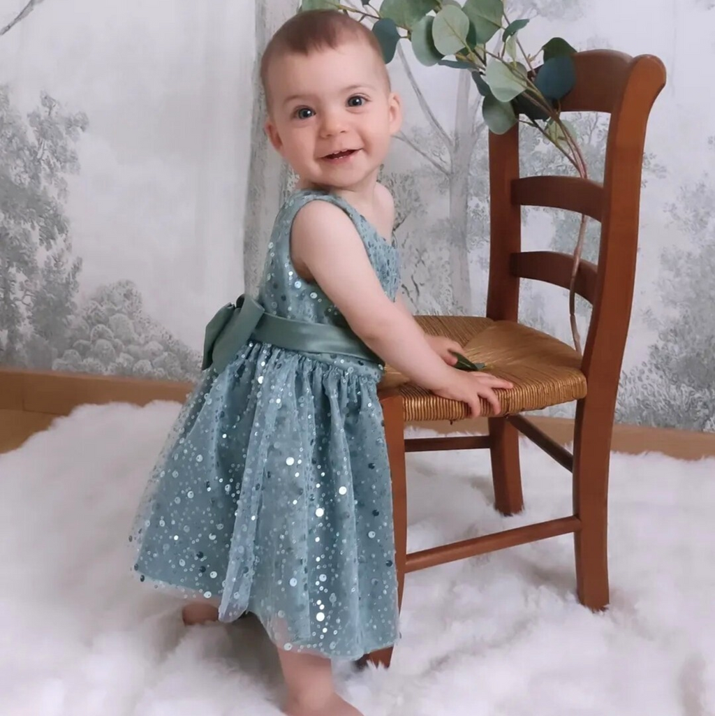 Petite fille souriante portant la robe de cérémonie vert sauge à sequins. Vue en pied montrant la coupe fluide et confortable idéale pour un mariage.