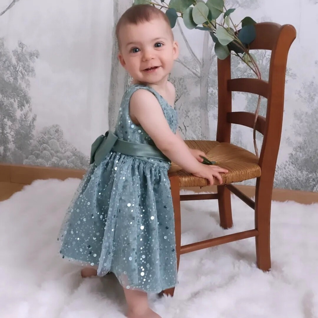 Petite fille souriante portant la robe de cérémonie vert sauge à sequins. Vue en pied montrant la coupe fluide et confortable idéale pour un mariage.