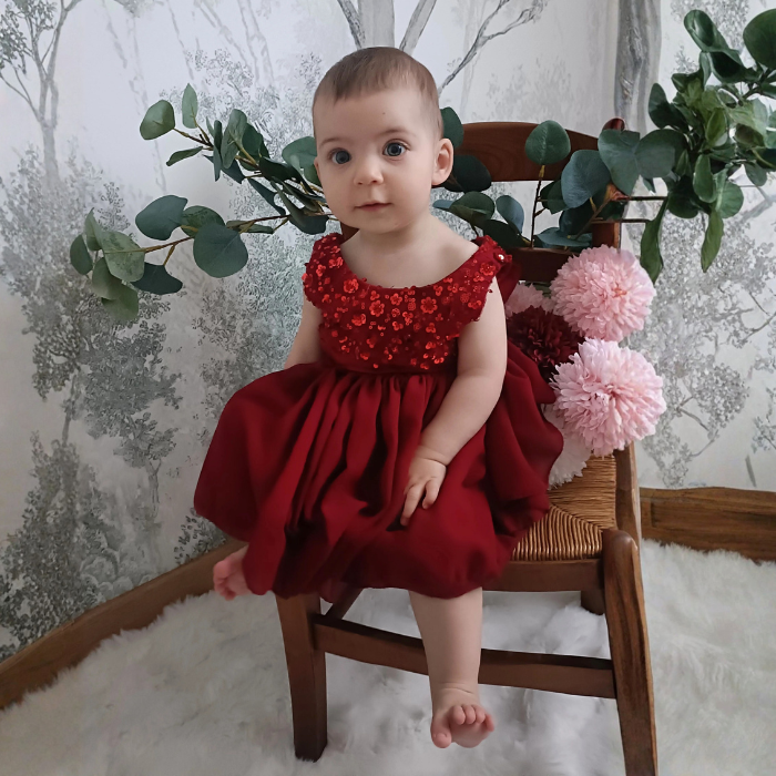 Bébé fille assise portant la robe de cérémonie rouge rubis à sequins. Vue de face montrant le confort du bustier et le volume de la jupe en tulle. Tenue de fête idéale.