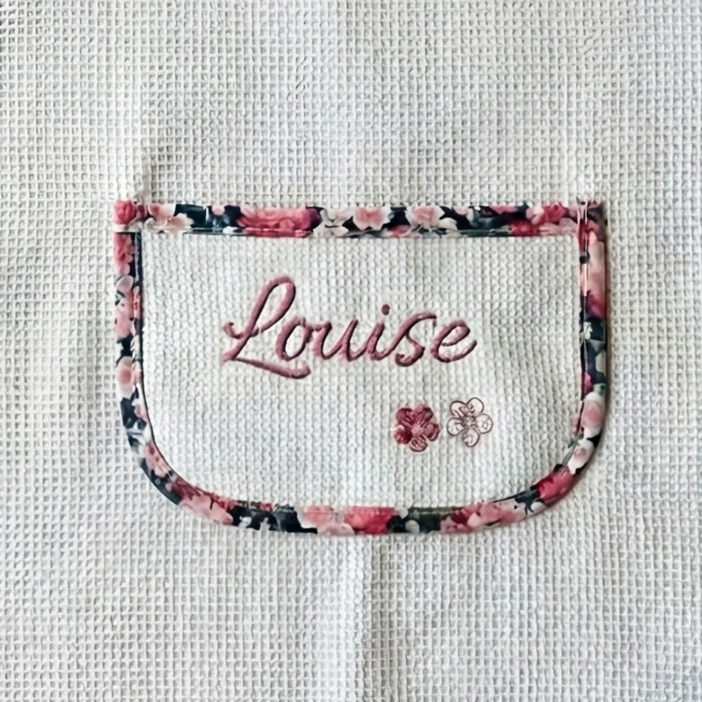Gros plan sur la poche ventrale du tablier enfant : broderie soignée du prénom Louise en rose et petites fleurs, sur tissu nid d'abeille blanc.