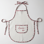 Gros plan sur la poche ventrale du tablier enfant : broderie soignée du prénom Louise en rose et petites fleurs, sur tissu nid d'abeille blanc.